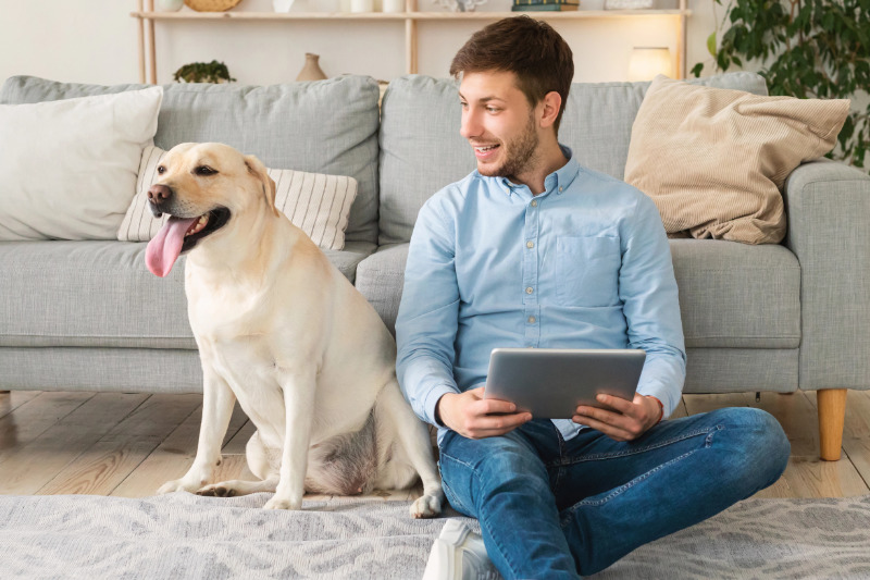 Mann sitzt mit seinem Labrador im Wohnzimmer auf dem Boden und schaut auf ein Tablet